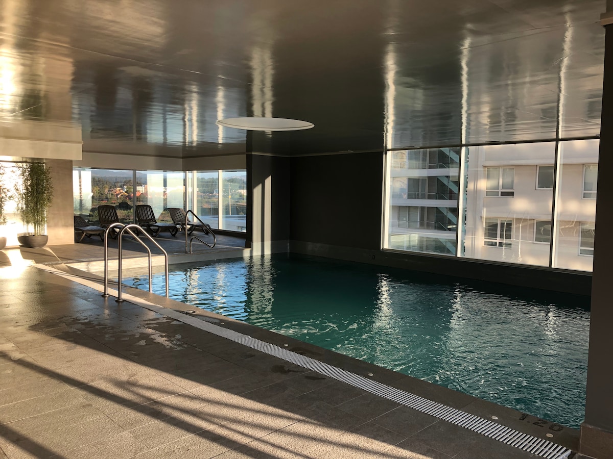An indoor pool area is highlighted by large windows that allow natural light to fill the space. The water is calm and inviting, with three lounge chairs positioned nearby. The clean lines and modern design of the surroundings create a serene atmosphere.