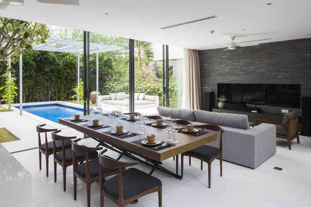 An open-plan living area features a modern dining table set for eight, with a view of the private swimming pool beyond large glass doors. A comfortable sofa sits nearby, and an entertainment unit is positioned against a stone-accent wall, enhancing the inviting space.