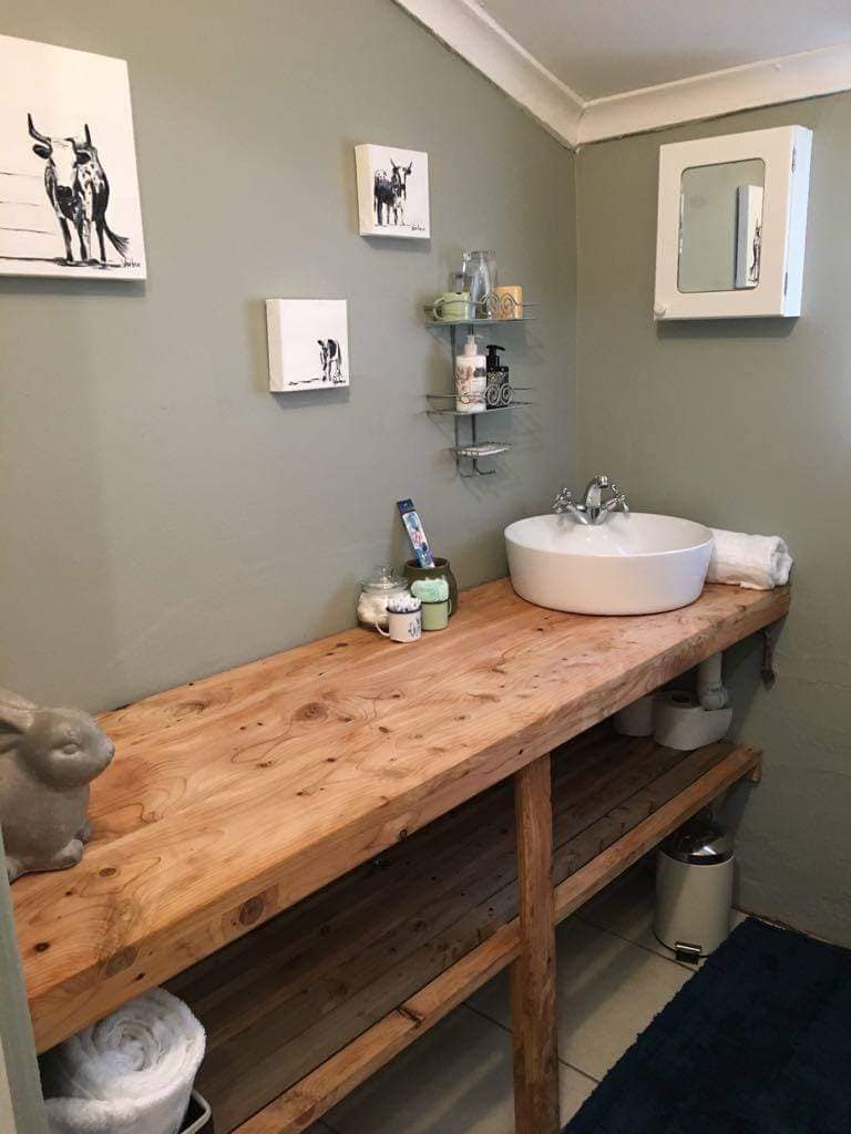 A wooden countertop features a round, white basin positioned on top, accompanied by an assortment of toiletries. A wall-mounted mirror is situated above the basin. Shelves display decorative items, while light-colored walls and tiled flooring create a clean and functional space.