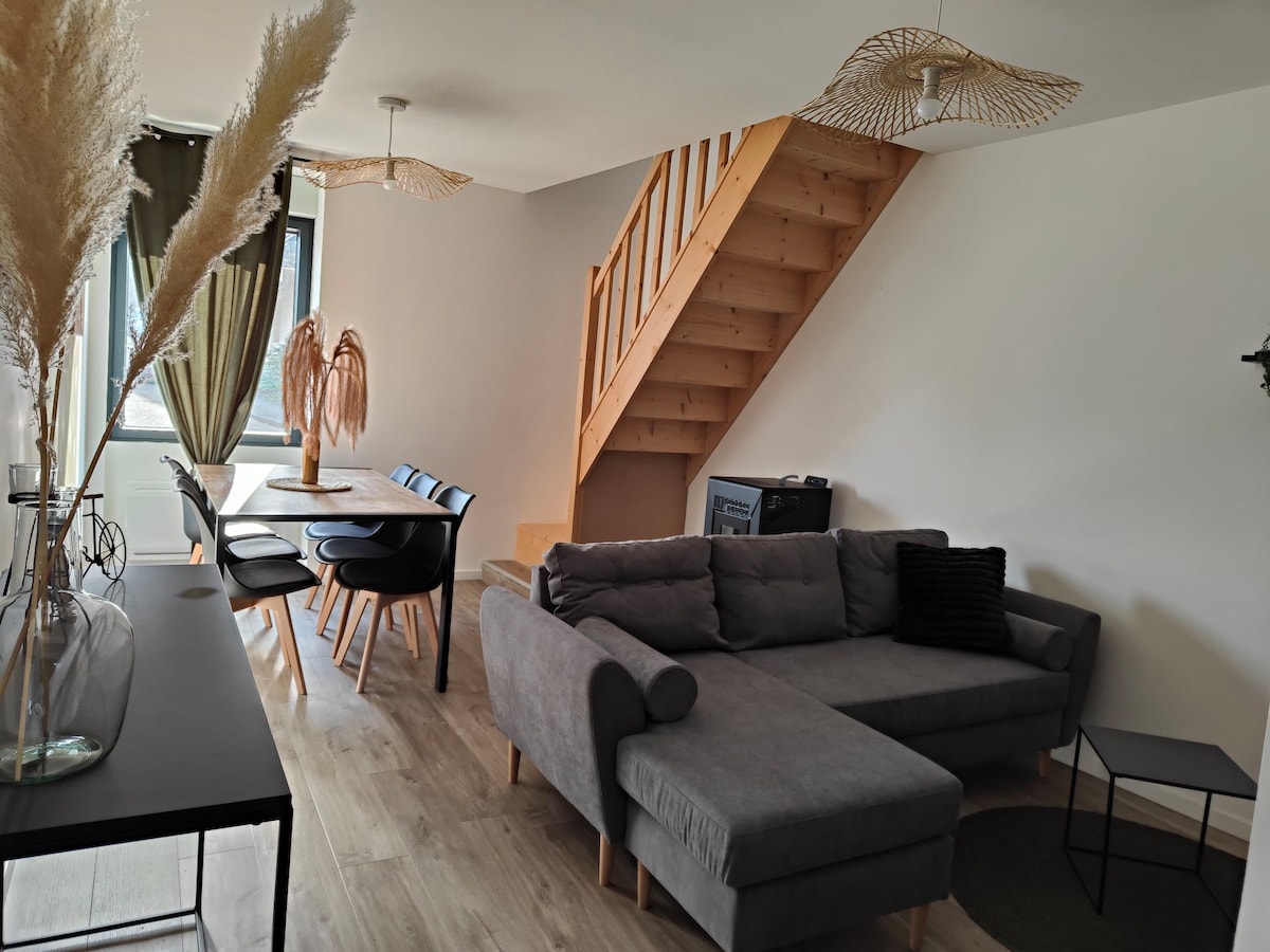 A spacious living area features a comfortable gray sectional sofa, a black coffee table, and a dining table with black chairs. Natural light pours in through large windows, with wooden stairs leading to the upper level. Decorative accents include pampas grass and woven light fixtures.