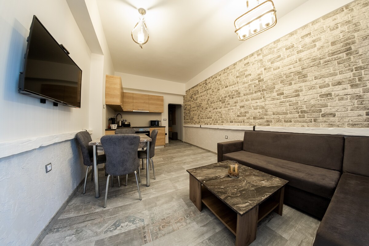 A spacious living area is depicted, featuring a sectional sofa and a low coffee table. A flat-screen TV is mounted on the wall opposite the seating. A dining table with four chairs is positioned nearby, enhancing the open layout. Natural light filters in through unseen windows.