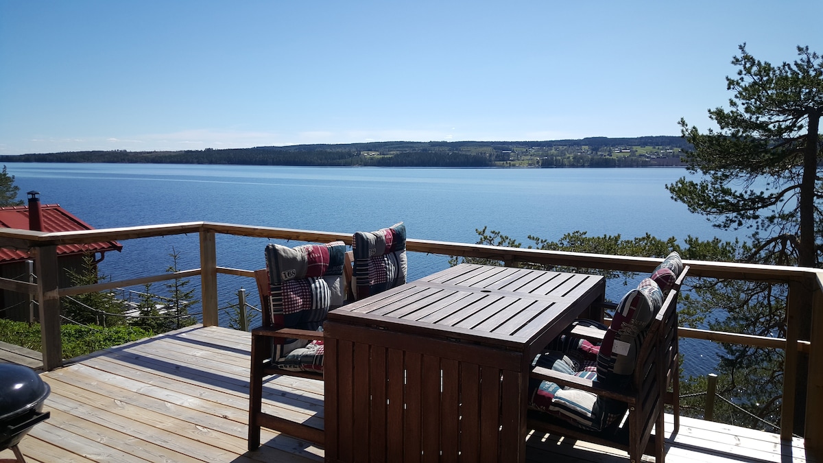 A spacious wooden deck features a large table surrounded by comfortable chairs, offering a clear view of the calm blue waters of the lake. Lush greenery can be seen in the distance, enhancing the serene atmosphere.