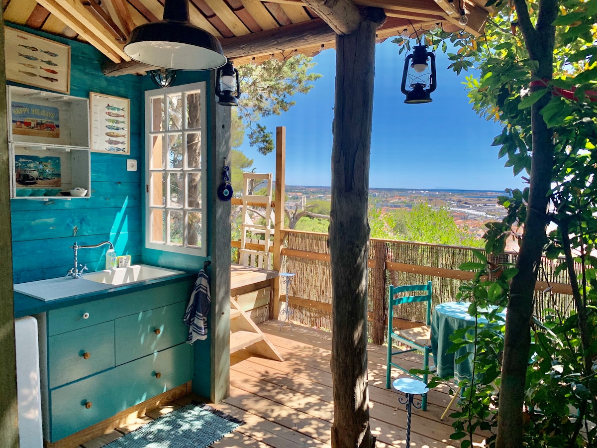 The image presents a cozy kitchen area with a teal color scheme, featuring a sink and exposed wooden beams. A small table and chair set on the terrace overlook scenic views of the city and harbor, framed by greenery and natural elements.
