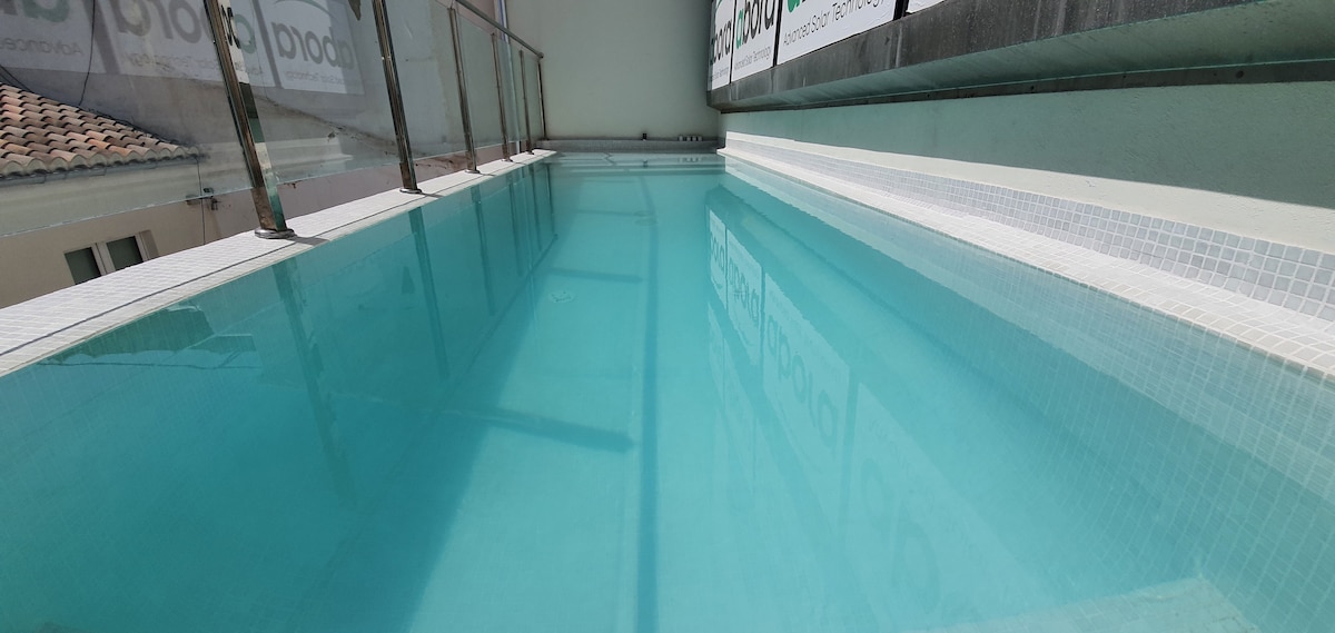 A tranquil swimming pool is shown, featuring clear, blue water and surrounded by a glass railing. The pool is situated on a terrace, emphasizing a serene atmosphere. Natural light reflects off the water, creating a calming visual effect.
