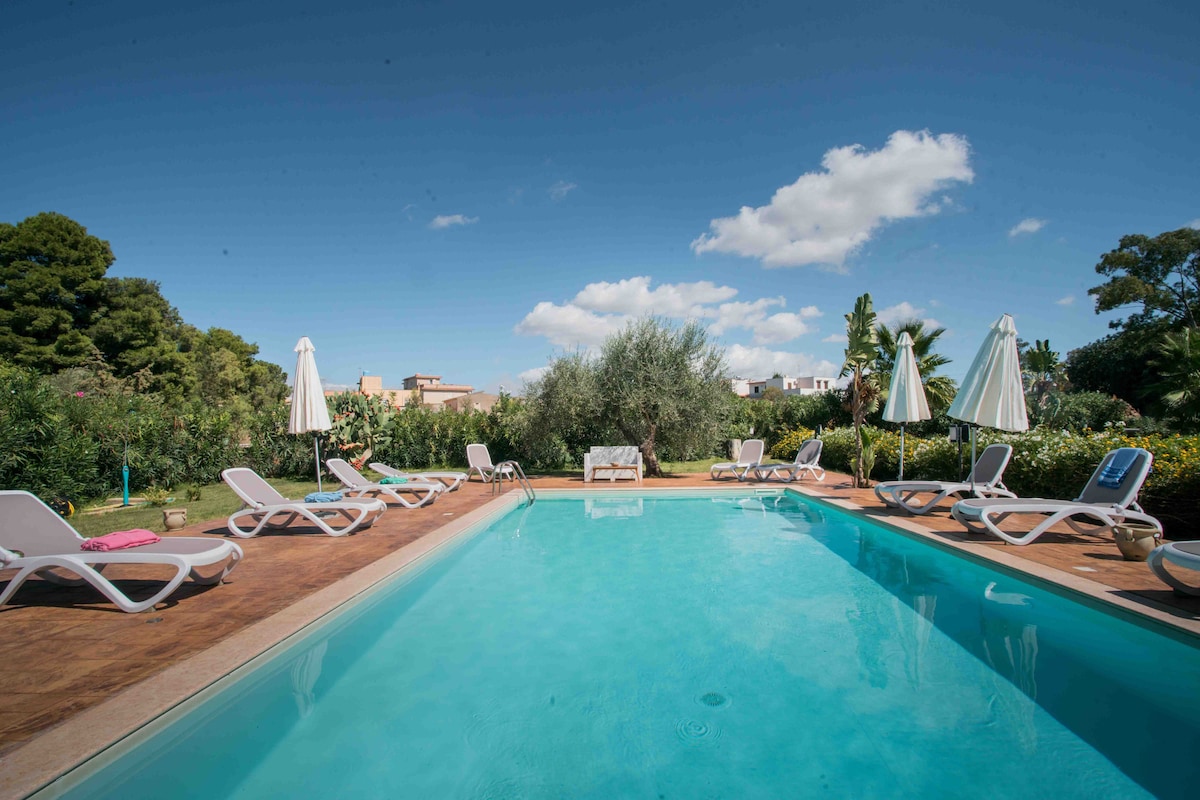 A clear blue swimming pool is surrounded by several lounge chairs and umbrellas. Lush greenery and flowering plants create a serene backdrop, with trees visible in the distance. The sky is bright and partly cloudy, contributing to a refreshing outdoor setting.