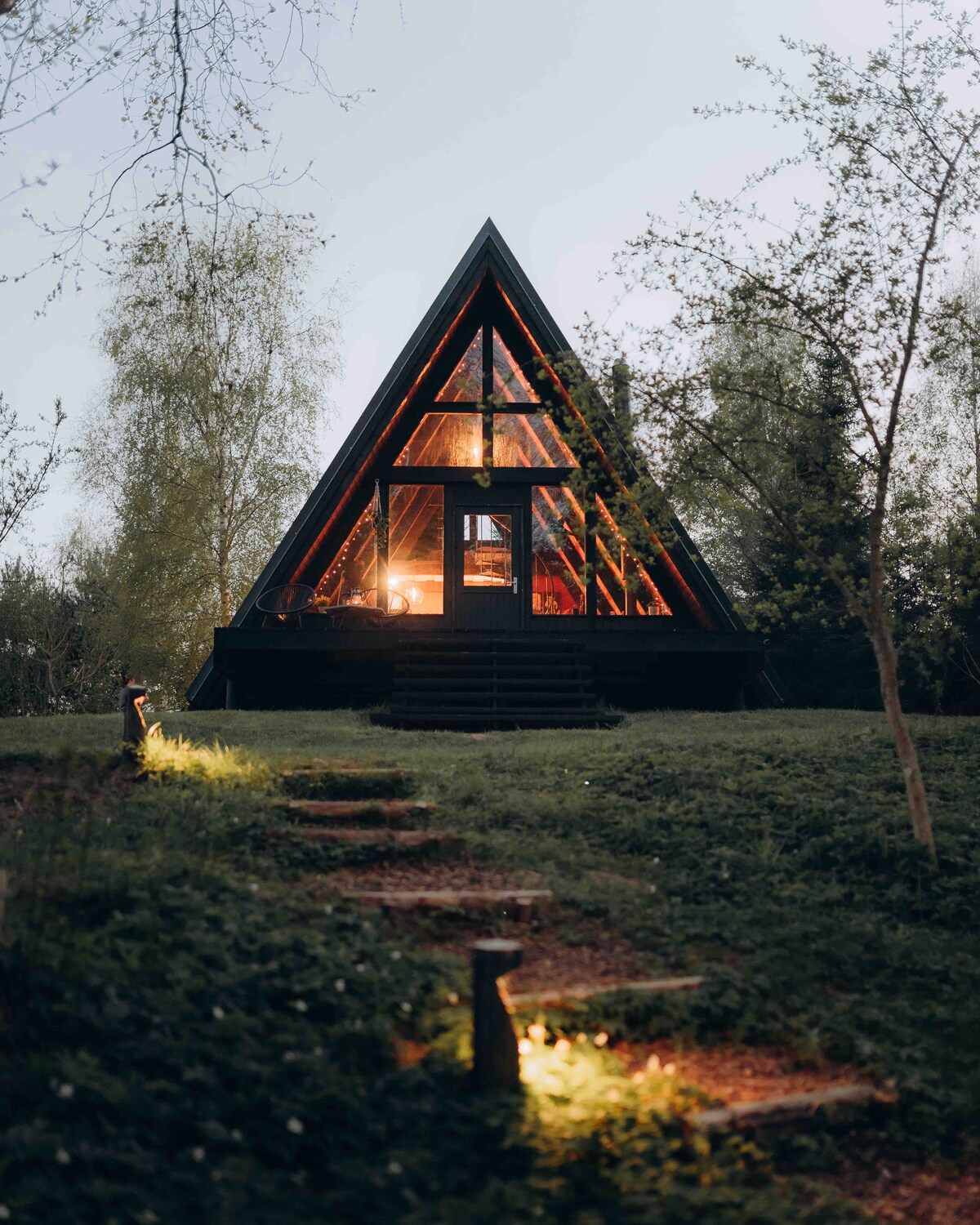 The A-frame house is nestled in a serene setting, with warm light glowing from its large windows, creating a cozy ambiance. Steps lead up to the entrance, surrounded by well-kept grass and trees. The structure's pointed roof enhances its unique aesthetic.