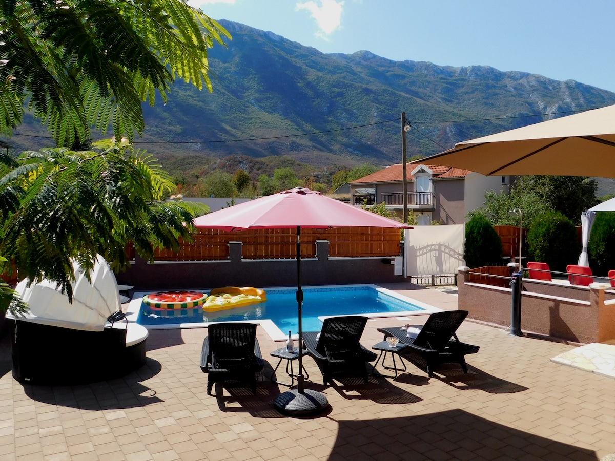 An outdoor area features a swimming pool surrounded by several lounge chairs, shaded by umbrellas. Colorful flotation devices are placed in the pool. In the background, the imposing Biokovo mountain range rises against a clear blue sky.