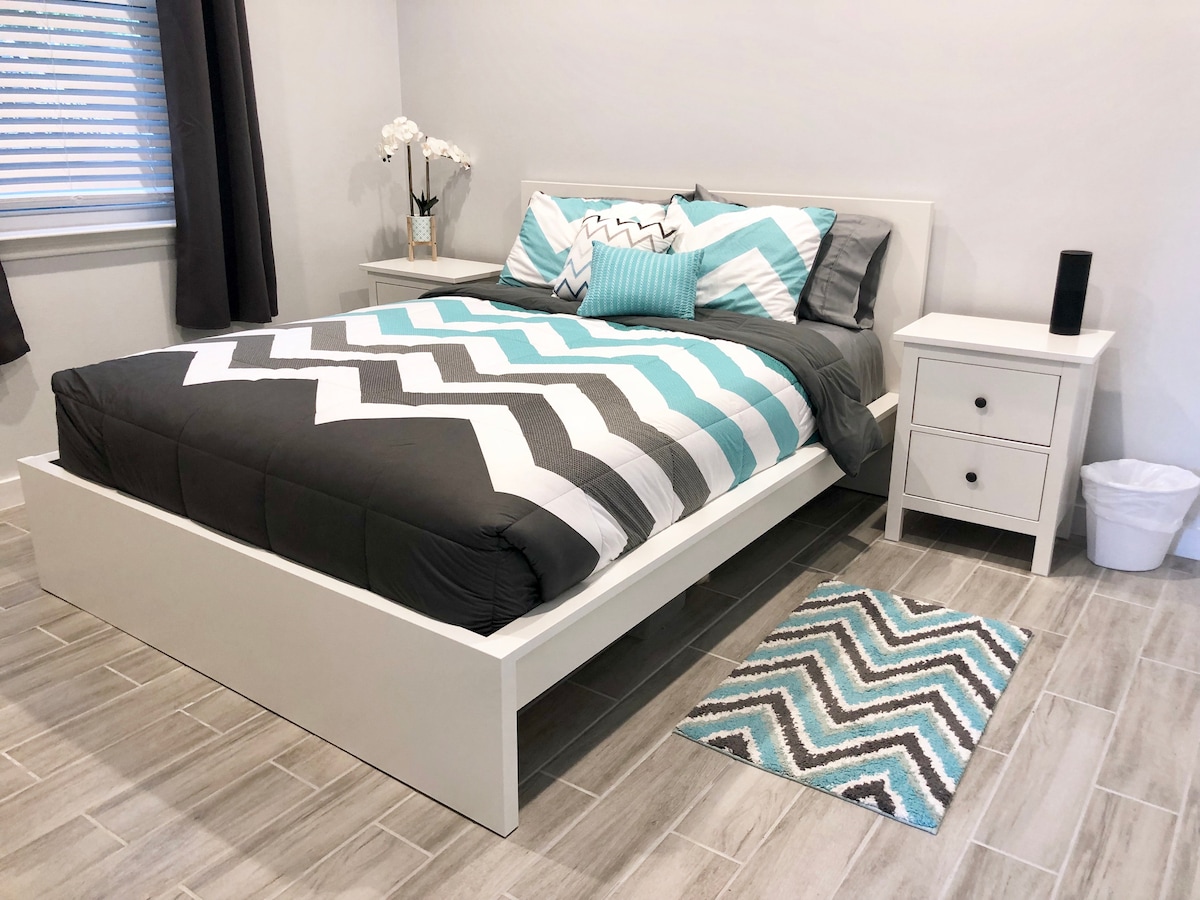 A well-appointed sleeping area features a modern queen bed with a patterned comforter in shades of grey and turquoise. A nightstand with a small lamp complements the bed, while a soft rug lays on the tiled floor, adding comfort to the space.