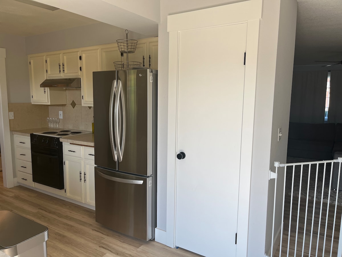 A modern kitchen area is shown, featuring stainless steel appliances including a refrigerator and oven. Light-colored cabinets complement the wood-style flooring. A doorway on the right leads to another room, while an open space can be seen in the background.