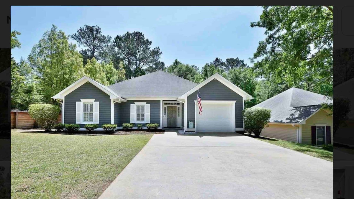 A single-story home features a gray exterior with white trim, set against a backdrop of lush greenery. A well-maintained front yard with small hedges frames the entrance. A driveway leads to the garage, offering easy access to the home.
