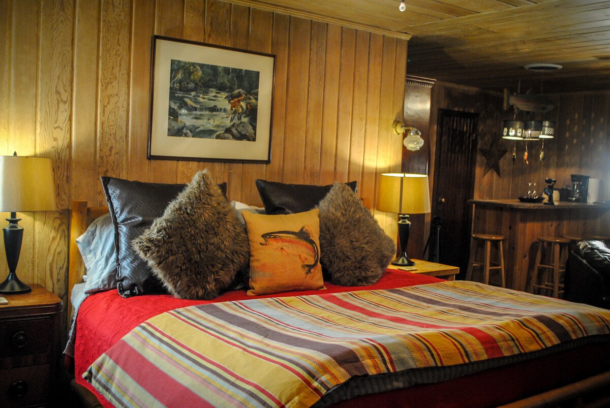 A cozy sleeping area is highlighted by warm wooden paneling and soft lighting. A spacious bed is adorned with a colorful striped blanket and plush pillows. A nature-themed artwork is displayed above the bed, and a small breakfast bar is visible in the background.