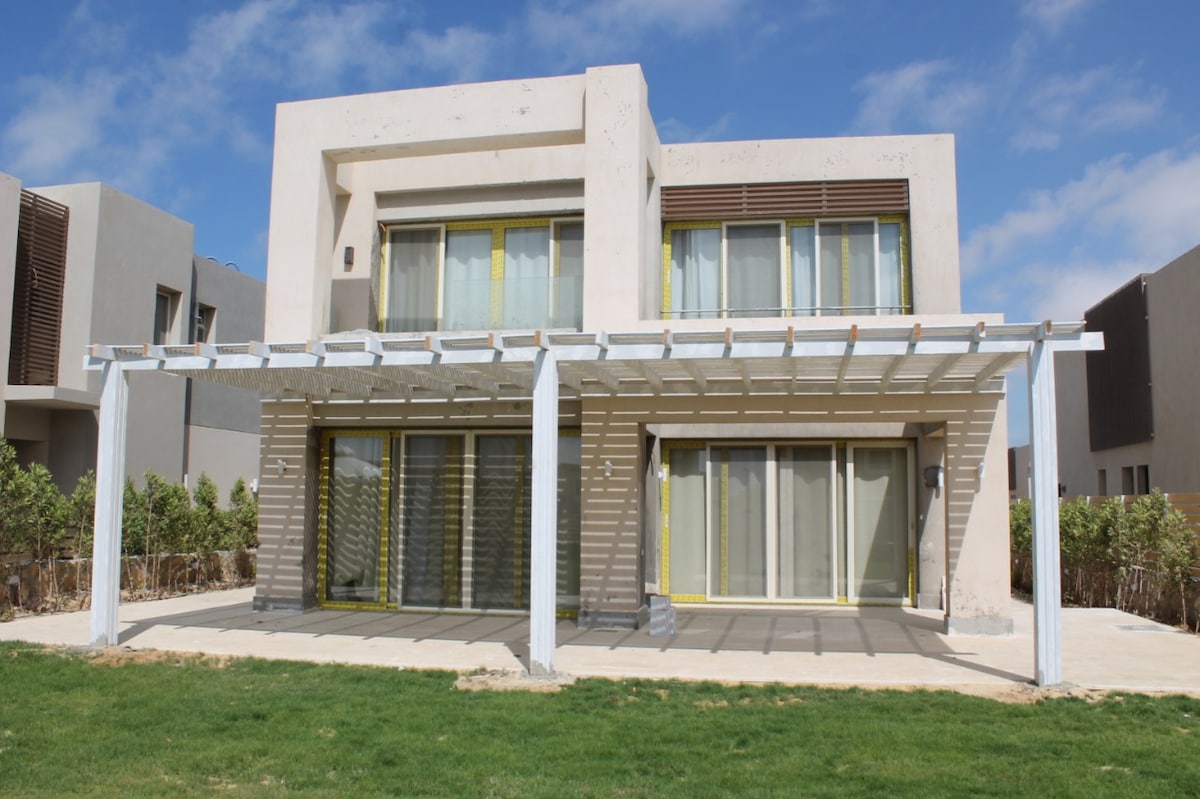 A modern villa is showcased with a well-maintained lawn and a spacious patio area. Large glass doors provide access to the interior, while a pergola extends overhead to create a shaded outdoor space. The building's contemporary design is complemented by a light-colored façade.
