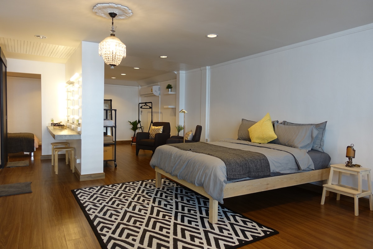 An open-plan room features a spacious bed with a grey duvet and an accent pillow, flanked by wooden side tables. A stylish area rug in geometric patterns covers the floor. A cozy seating area is present with armchairs, and a large mirror with bright lighting is positioned next to a wardrobe.