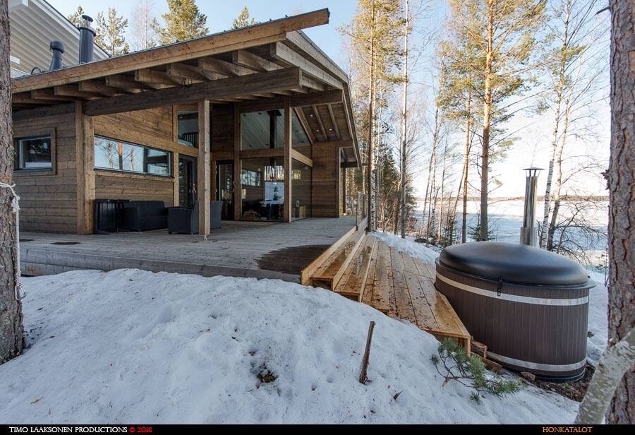 The exterior features a wooden deck that leads to a hot tub surrounded by snow and slender trees. Large glass walls showcase the inviting interior of the building, while the tranquil lake is visible in the background, reflecting the natural landscape.