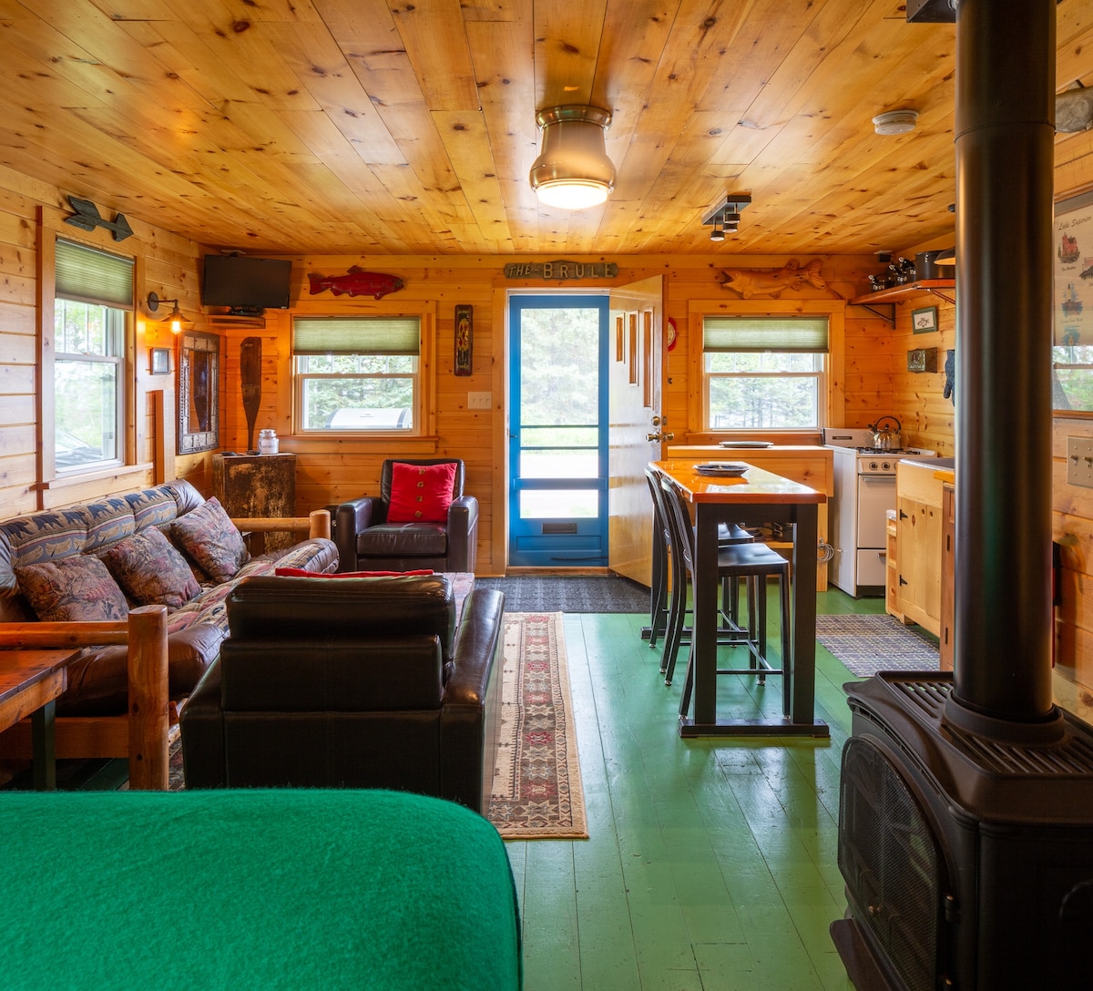 The interior of the cabin is characterized by warm wood paneling and a spacious layout. A mix of seating arrangements, including a cozy couch and armchair, is complemented by a dining table and kitchen area. Large windows provide natural light, and a wood stove adds a rustic touch.