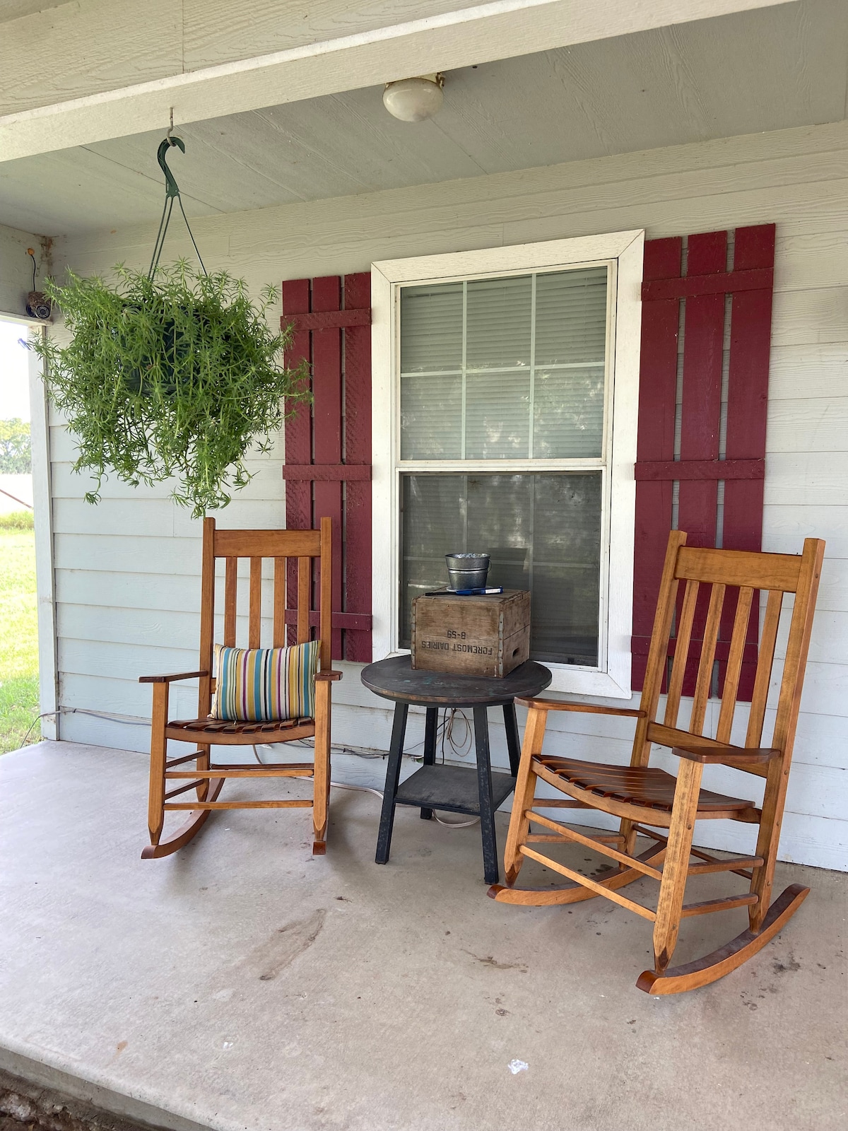Two wooden rocking chairs are positioned on a covered porch, accompanied by a small table. A hanging plant basket adds greenery above, while a striped pillow rests on one chair. A window with red shutters is visible in the background.