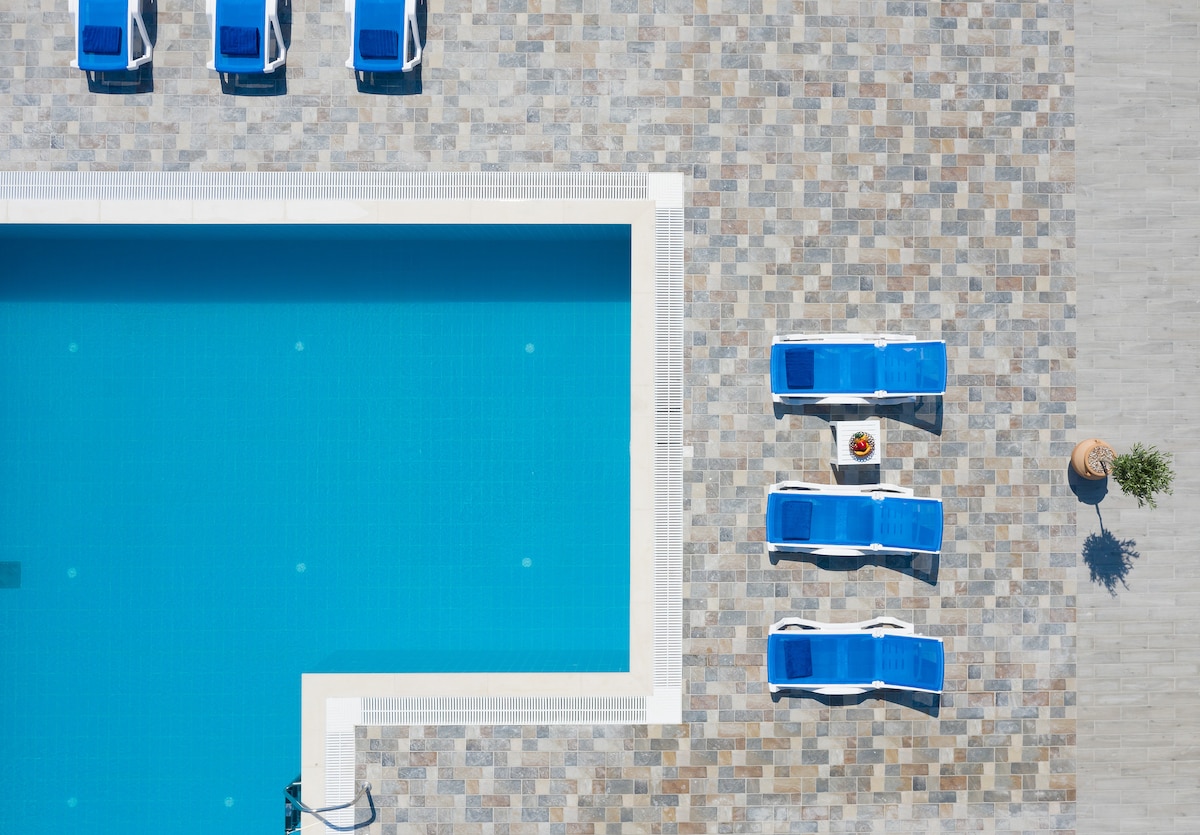 An aerial view reveals a large private pool surrounded by a stone patio. Blue sunbeds with white frames are placed around the pool, alongside a small round table with a potted plant. The clear blue water reflects sunlight, enhancing the serene setting.