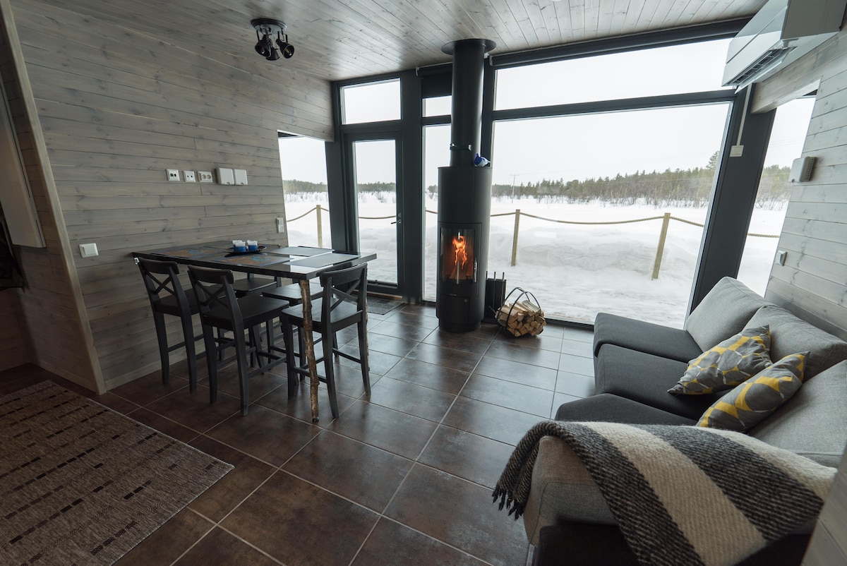 A cozy living area is visible, featuring a simple sofa adorned with patterned cushions. A dining table with several chairs faces expansive glass walls, allowing views of the snowy landscape outside. A modern fireplace stands in the corner, enhancing the warmth of the space.