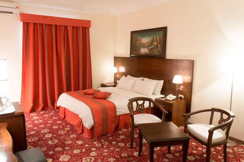 A well-appointed bedroom features a spacious bed dressed in neutral linens and accented with decorative pillows. A rich red area rug covers the floor, and a painting hangs above the headboard. Two wooden chairs and a small table are placed beside the bed.