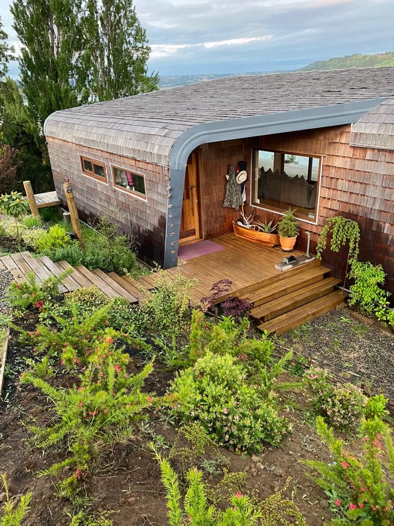 A unique house design is showcased with a curved roof and wooden exterior. Steps lead to the entrance, surrounded by low greenery and a wooden walkway. Large windows reflect the natural light, contributing to the welcoming appearance of the home.