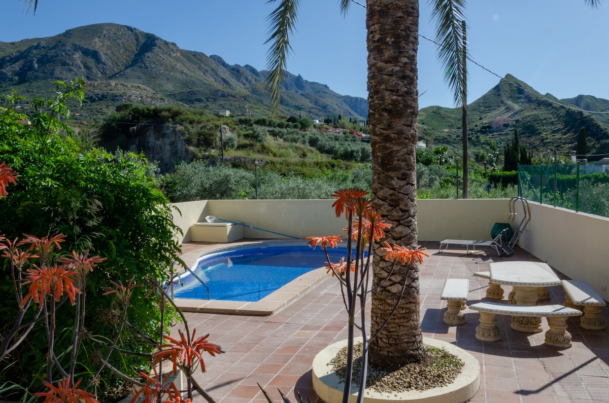A tranquil outdoor space features a private pool surrounded by mountains. Colorful plants frame the area, and a stone table sits nearby, providing a place to relax. The sunlit landscape creates a serene atmosphere for outdoor enjoyment.