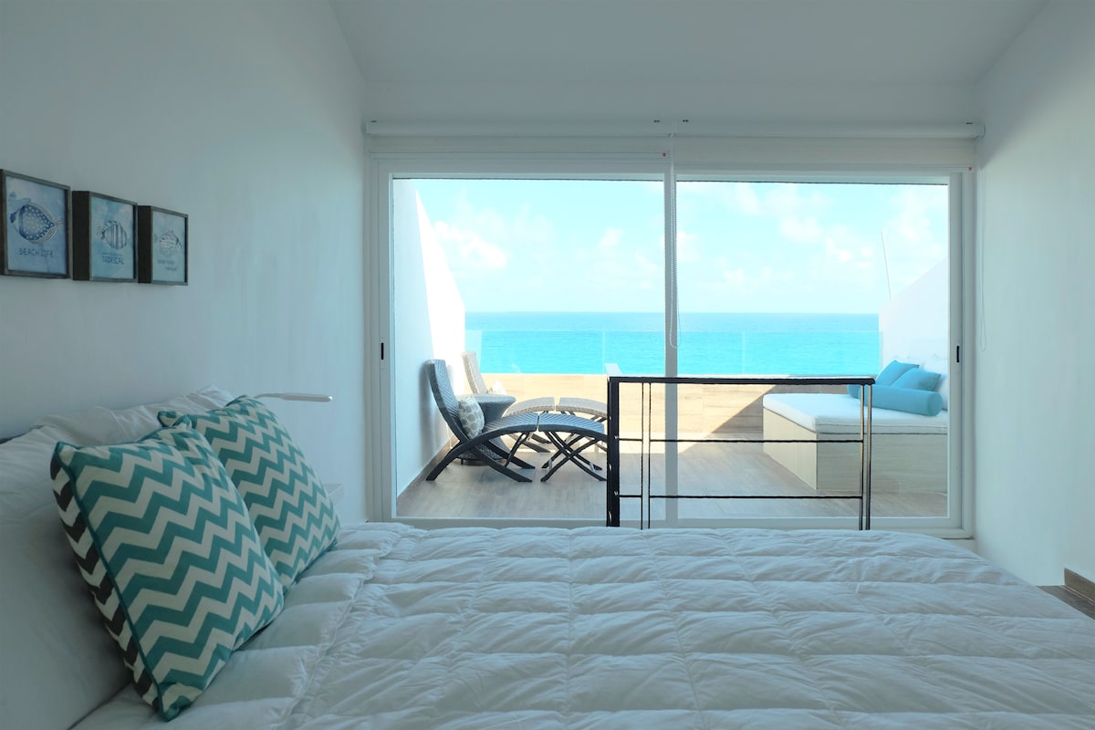 A well-appointed bedroom features a spacious bed adorned with patterned pillows. A large glass door opens to a terrace with ocean views, where lounge chairs are positioned to enjoy the scenery. Soft natural light floods the room, creating a serene atmosphere.