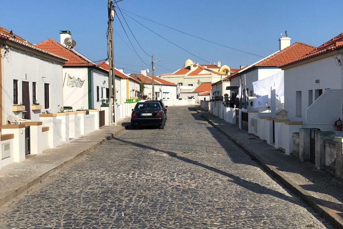 A quiet, cobblestone street is lined with charming white and pastel-colored houses, featuring terracotta roofs. A parked car is visible on one side, while light blue skies create a serene atmosphere. The simplicity of the surroundings reflects the area's tranquil charm.