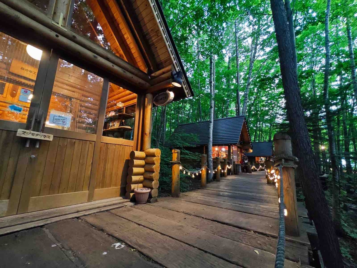 A pathway is shown leading through trees, illuminated by soft lights along the sides. Wooden structures can be seen in the distance, featuring a blend of natural materials and rustic design elements. The surrounding foliage creates a serene and inviting atmosphere.