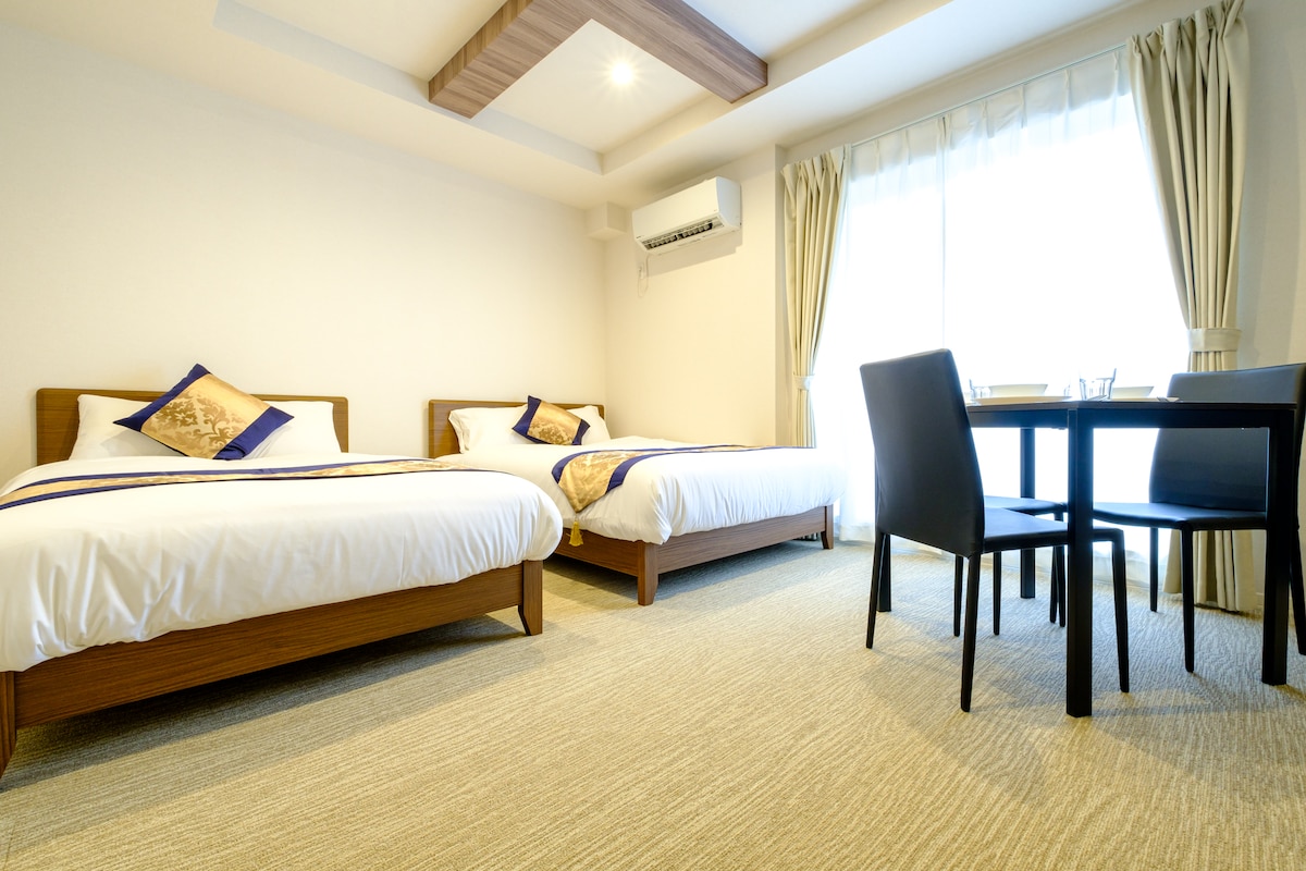 A well-lit room featuring two spacious beds with decorative pillows. A small table and chairs are positioned beside the large window, which is dressed with light curtains, allowing natural light to brighten the space. The room is adorned with neutral tones and a clean design.