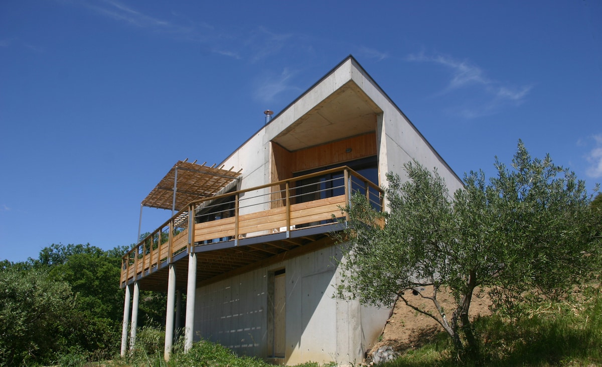 The modern triangular building features a wooden terrace with a railing, elevated on sturdy pillars. The exterior showcases a combination of raw concrete and warm timber elements. Surrounding greenery, including olive trees, enhances the serene setting against a clear blue sky.