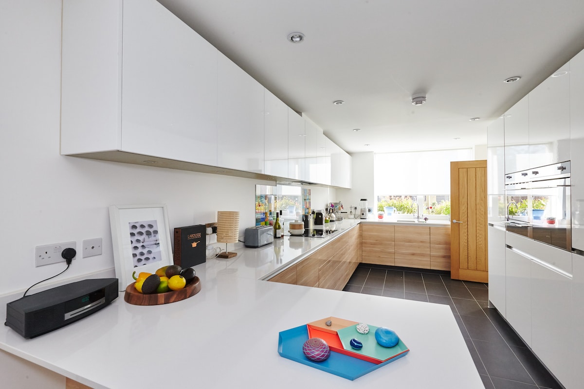 A modern kitchen features sleek white cabinetry and an extensive countertop. Natural light fills the space, highlighting a wooden accent wall. An assortment of fruits is artistically arranged on a wooden platter, complemented by colorful decorative items placed on a vibrant tray.