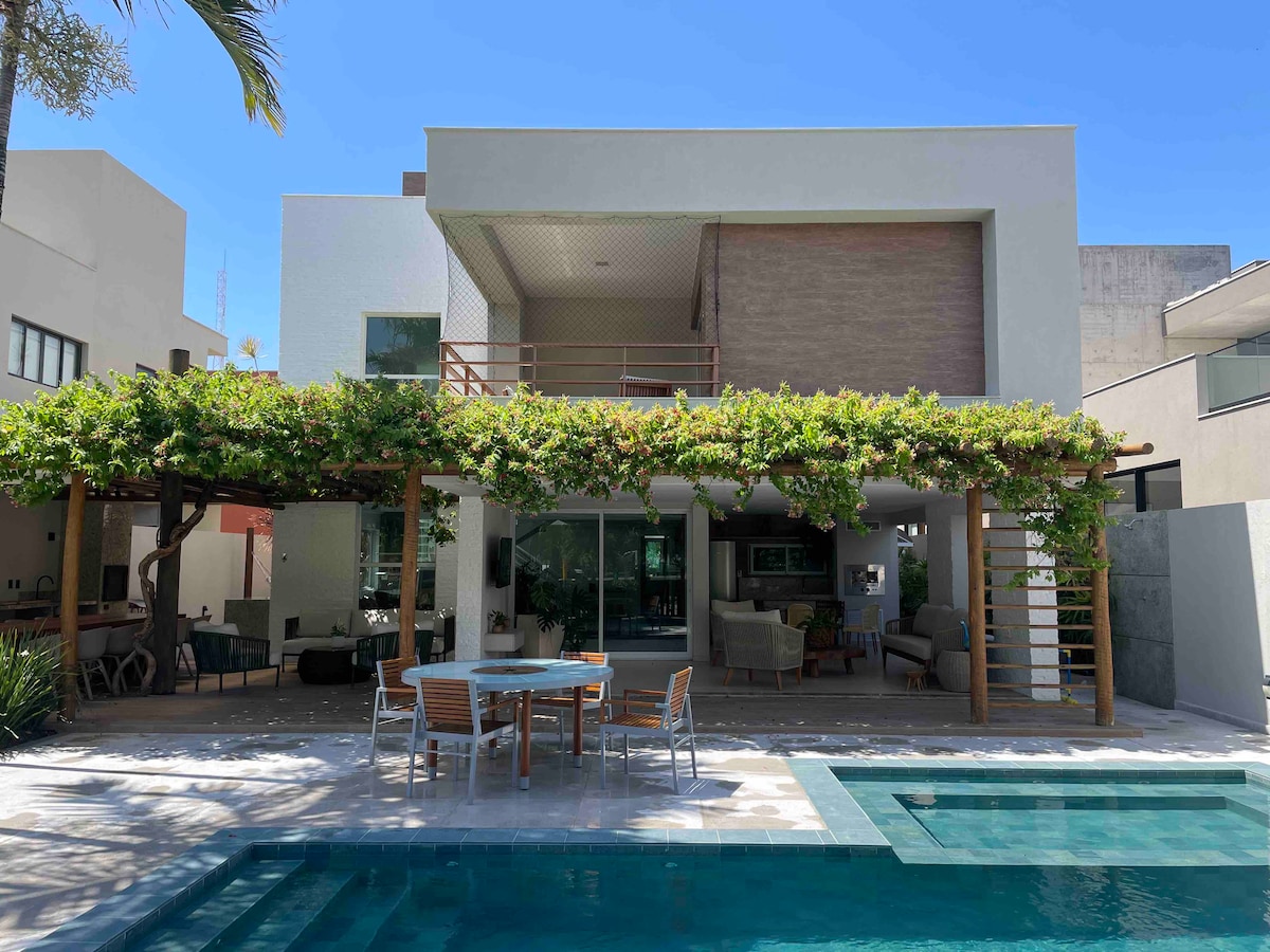 A spacious outdoor area is featured, showcasing a beautifully landscaped patio with a swimming pool. The house displays a modern two-story design, with large windows allowing natural light. A covered dining space is adorned with greenery, offering a relaxing atmosphere for outdoor gatherings.