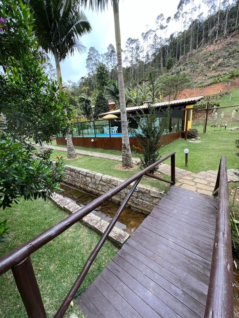 A wooden bridge leads to a grassy area surrounded by palm trees. A stream flows alongside the pathway, with a secluded swimming pool visible in the distance, bordered by vibrant plants and trees in the background.