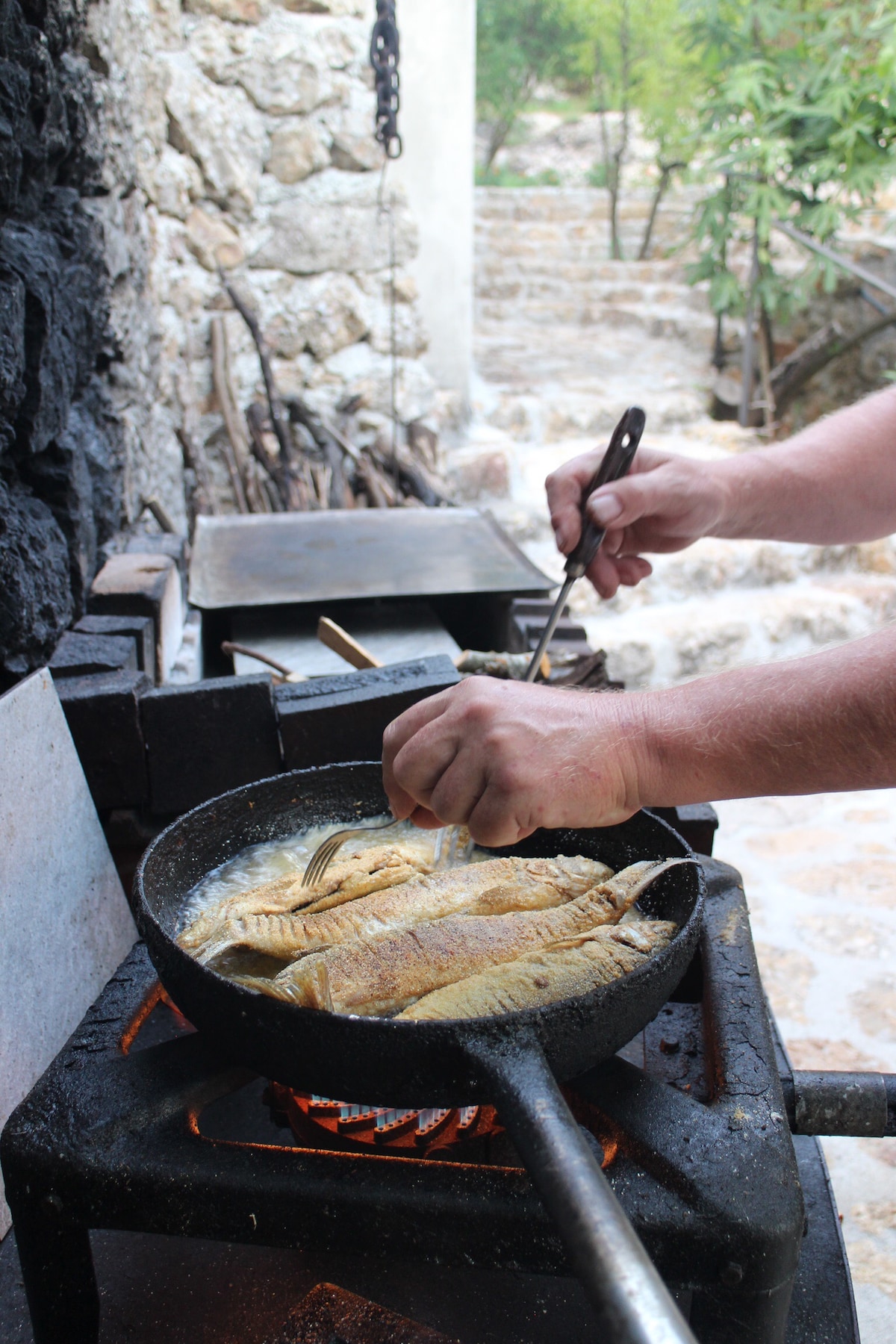 A cast iron skillet is placed on a stove, with fillets being flipped using tongs. The backdrop features a stone wall and visible steps leading to a garden area, surrounded by greenery. Natural light enhances the cooking space.