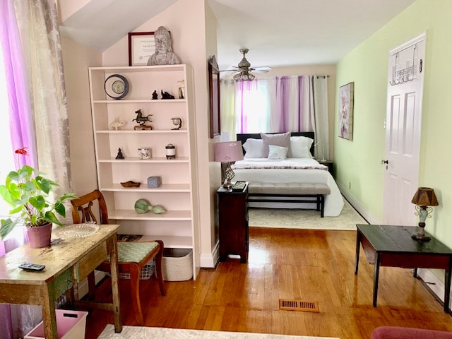 The room features a cozy layout with a spacious bed adorned by light-colored linens. A small bookshelf displays decorative items and a plant, while two tables provide space for relaxation. Natural light filters through sheer curtains, creating a calming atmosphere.
