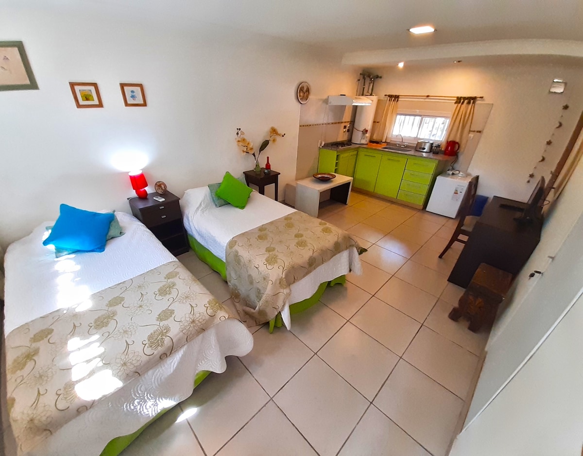A modern studio features two spacious beds adorned with light decorative bedspreads. A bright green kitchen area offers ample storage, while natural light streams through a window, illuminating the open layout. A dining table and chairs are positioned adjacent to the kitchen.