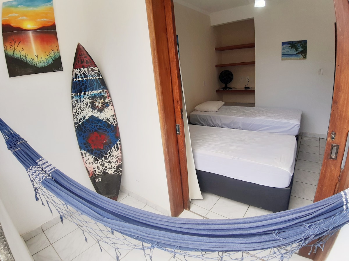An airy room features two spacious beds, one atop a dark base. A colorful surfboard is displayed against the wall, while a blue hammock hangs in the foreground. Art depicting a sunset is mounted above a shelf, enhancing the vibrant atmosphere.