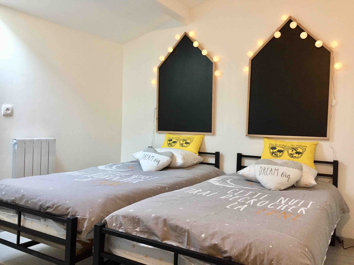 The room features two twin beds arranged side by side, each with decorative pillows showcasing playful designs. Warm string lights hang above, and the walls are painted in a light color, enhancing the cozy atmosphere. A radiator is positioned nearby for comfort.