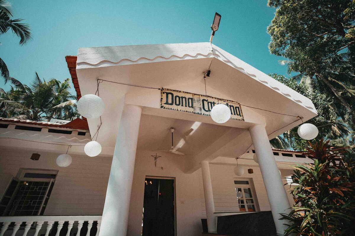 The guesthouse entrance features a charming white facade with a sloped roof and decorative tiles. Soft, round pendant lights hang from the overhang. Lush greenery and palm trees frame the structure, creating a serene atmosphere.