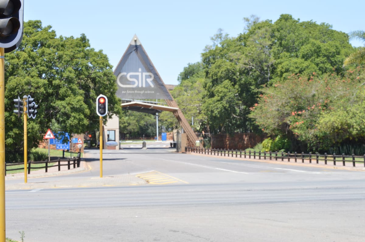 The entrance to the CSIR is prominently displayed, featuring a triangular structure with the acronym 'CSIR' visible. Lush green trees line the road, providing a natural backdrop. Traffic lights and road signs are present, indicating vehicle access to the area.