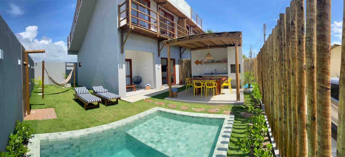 An outdoor space features a refreshing pool surrounded by lush greenery. A patio area includes a dining table with bright yellow chairs and a barbecue grill. Two sun loungers are set nearby, offering a relaxing atmosphere. The structure has a spacious upper terrace with wooden railings.