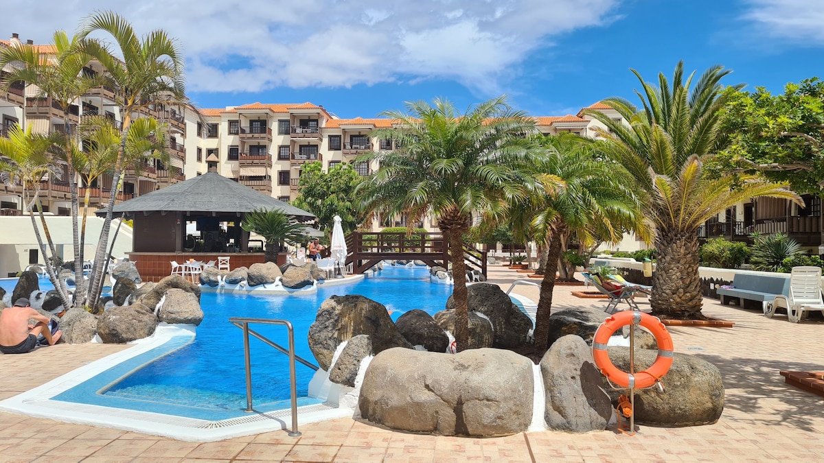A communal swimming pool is surrounded by lush palm trees and decorative rocks. Comfortable lounge chairs are arranged along the poolside, and a shaded seating area is visible under a thatched roof. The clear blue sky adds a bright backdrop to the tranquil setting.