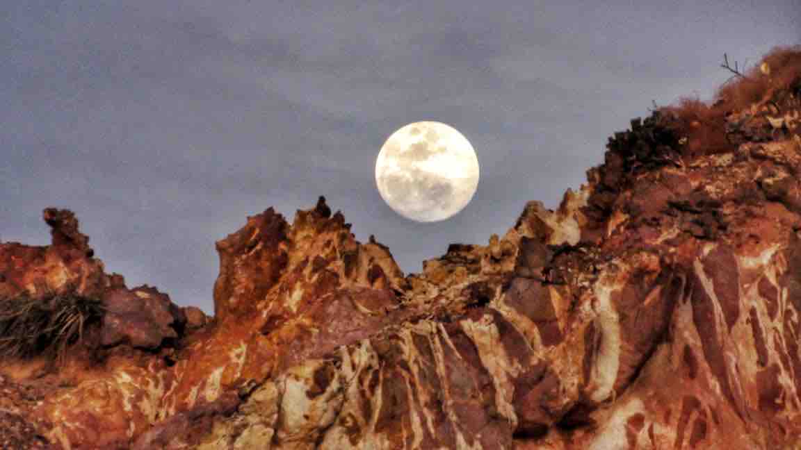 A large full moon is positioned above rugged, rocky terrain, displaying a variety of warm earth tones. The moon's surface features are visible, contrasting against the textured cliffs illuminated by the moonlight under a softly lit sky.
