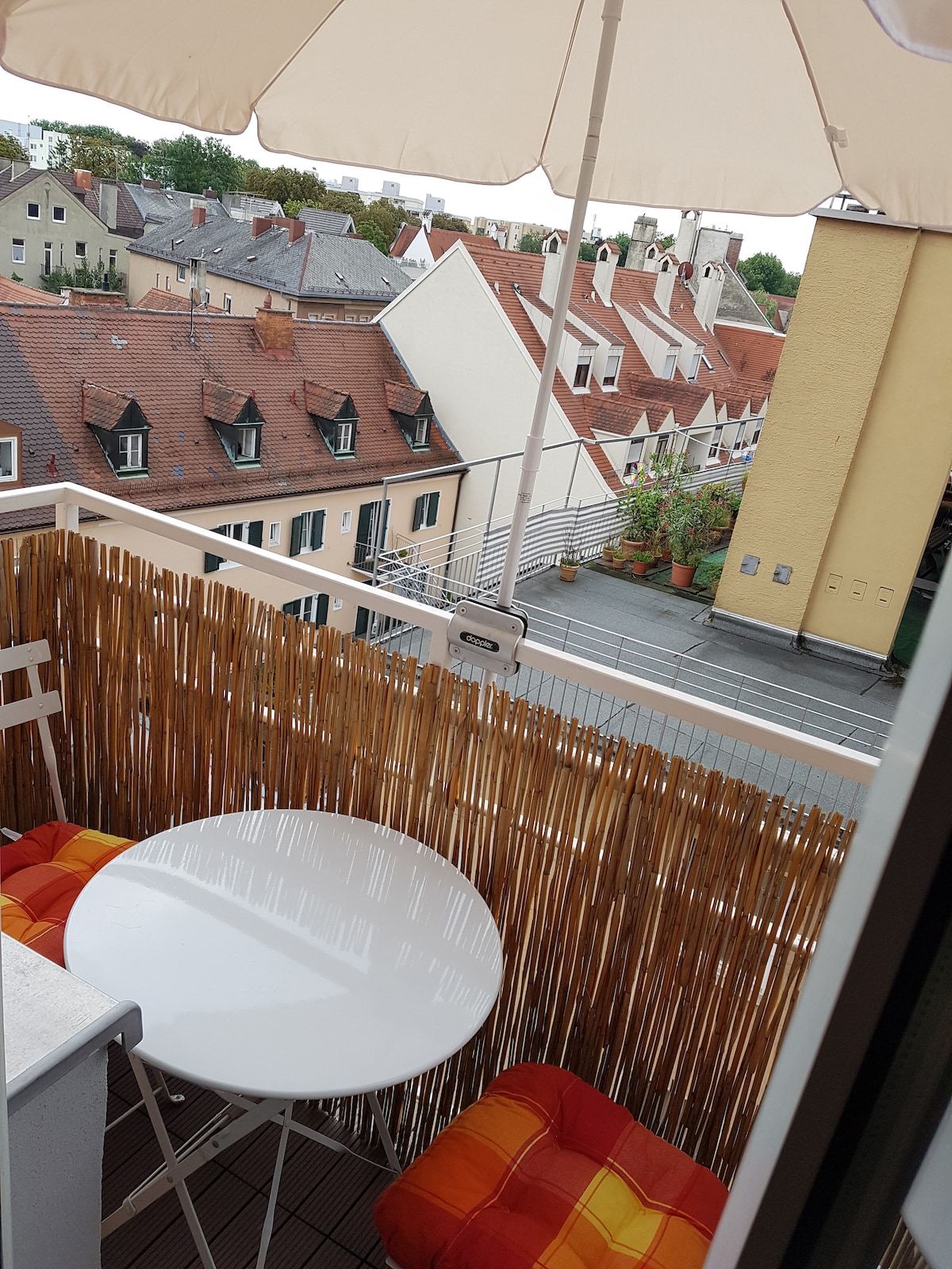 A small balcony is visible with a round white table and two cushioned chairs. A sun umbrella provides shade, while a bamboo screen offers privacy. Charming rooftops and buildings are seen in the background, creating a quaint urban view.