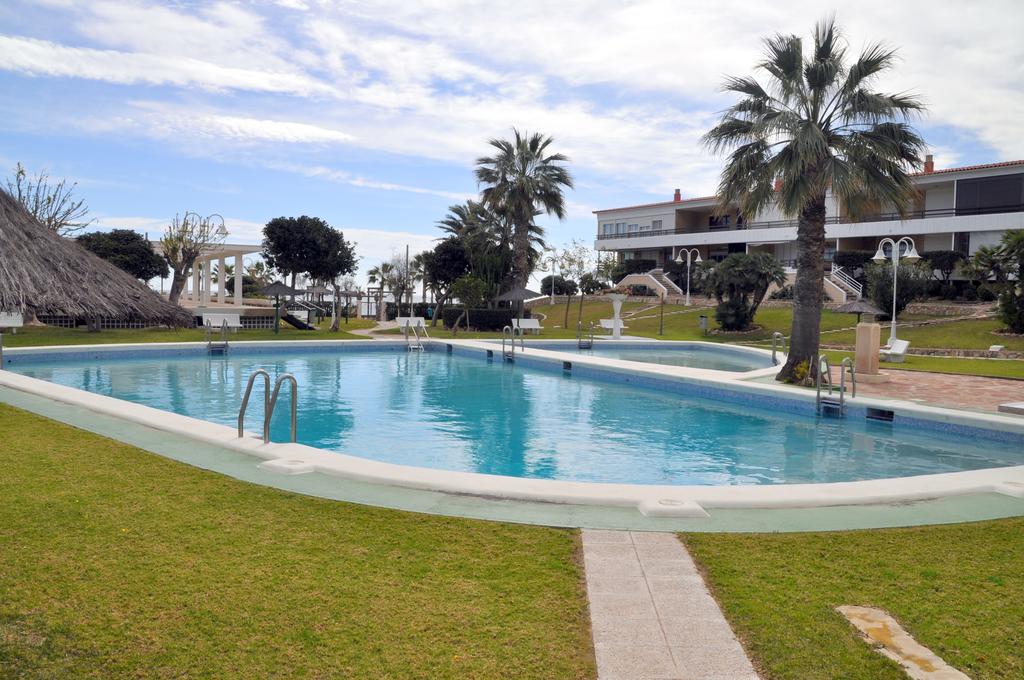 A clear swimming pool is surrounded by well-maintained grass and palm trees. Sun loungers are positioned alongside the pool, and a thatched-roof gazebo adds a tropical touch. The tranquil environment is complemented by landscaped gardens and a modern building visible in the background.