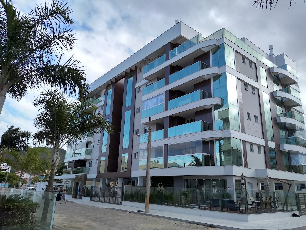 A modern multi-story building is depicted, featuring large glass windows that reflect the surrounding landscape. The exterior is complemented by balconies and palm trees in the foreground, creating an inviting ambiance. A clean and spacious area surrounds the entrance, enhancing accessibility.