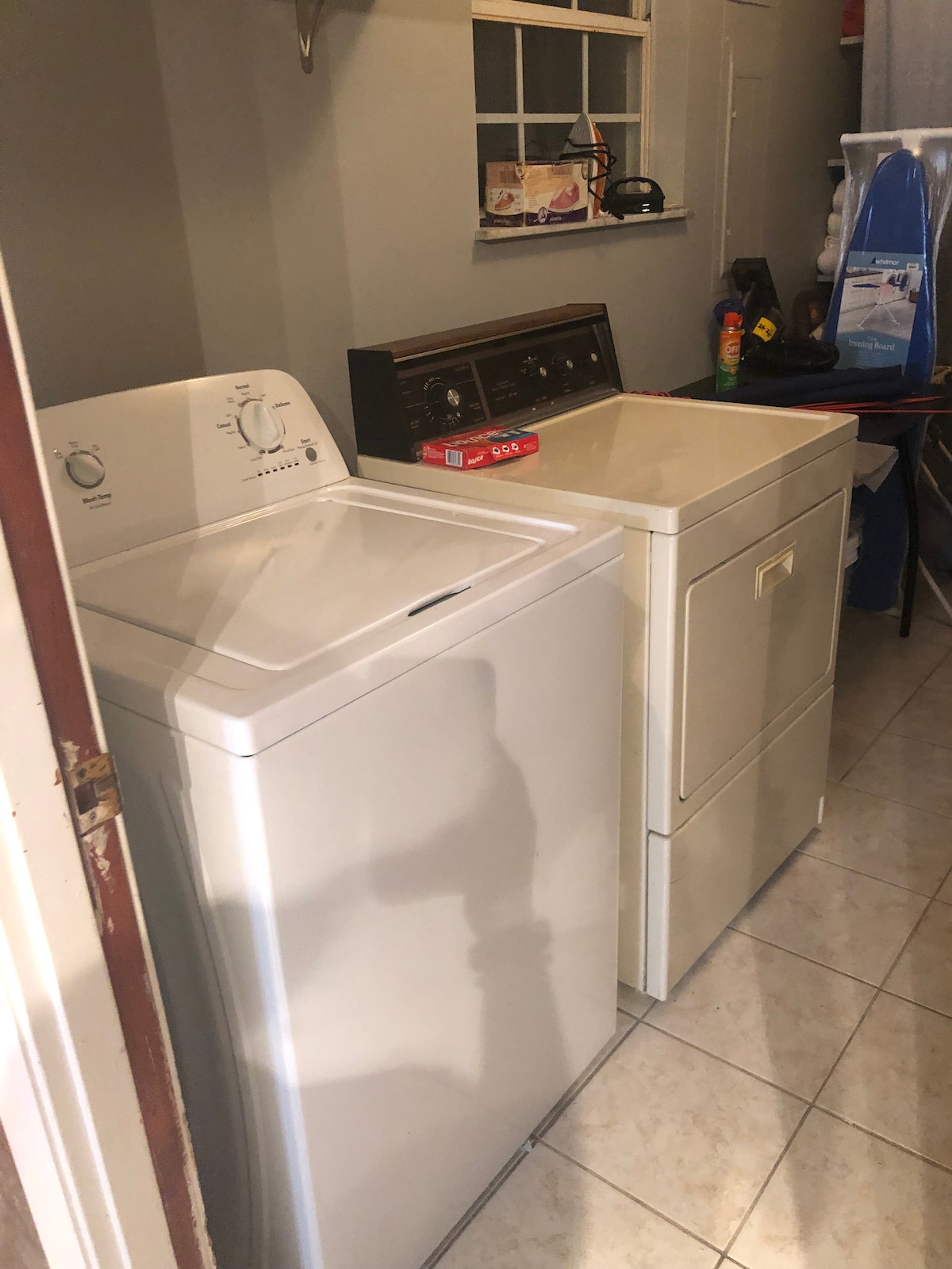 A compact laundry area features a white top-loading washing machine and a matching dryer, both situated on tiled flooring. Various cleaning supplies and household items are stored nearby, organized on shelves and in bins.