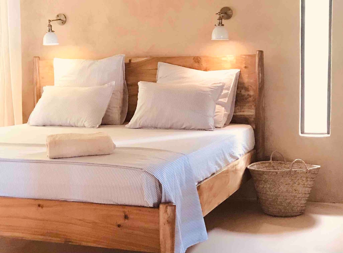 A spacious wooden bed is adorned with multiple pillows, accompanied by a neatly placed towel. Soft bedside lighting highlights the rustic charm of the natural wood. A woven basket is positioned beside the bed, while a narrow window provides a glimpse of natural light.
