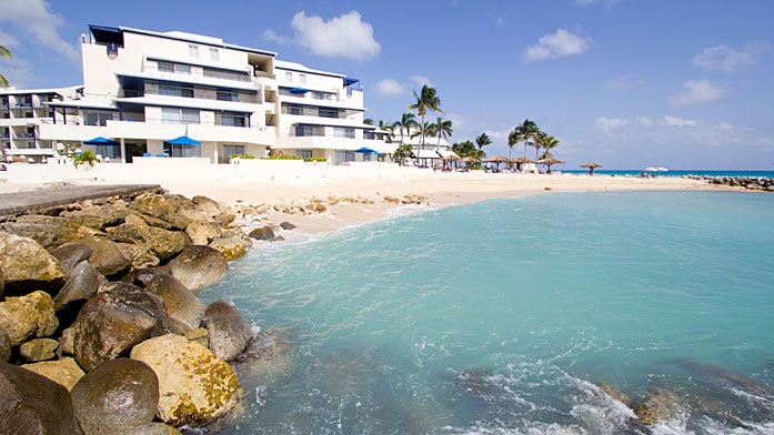 A beachfront view captures the resort's modern architecture and lush palm trees. The clear turquoise waters gently lap against rocky outcrops, while soft white sand lines the shore, inviting guests to enjoy the seaside setting.