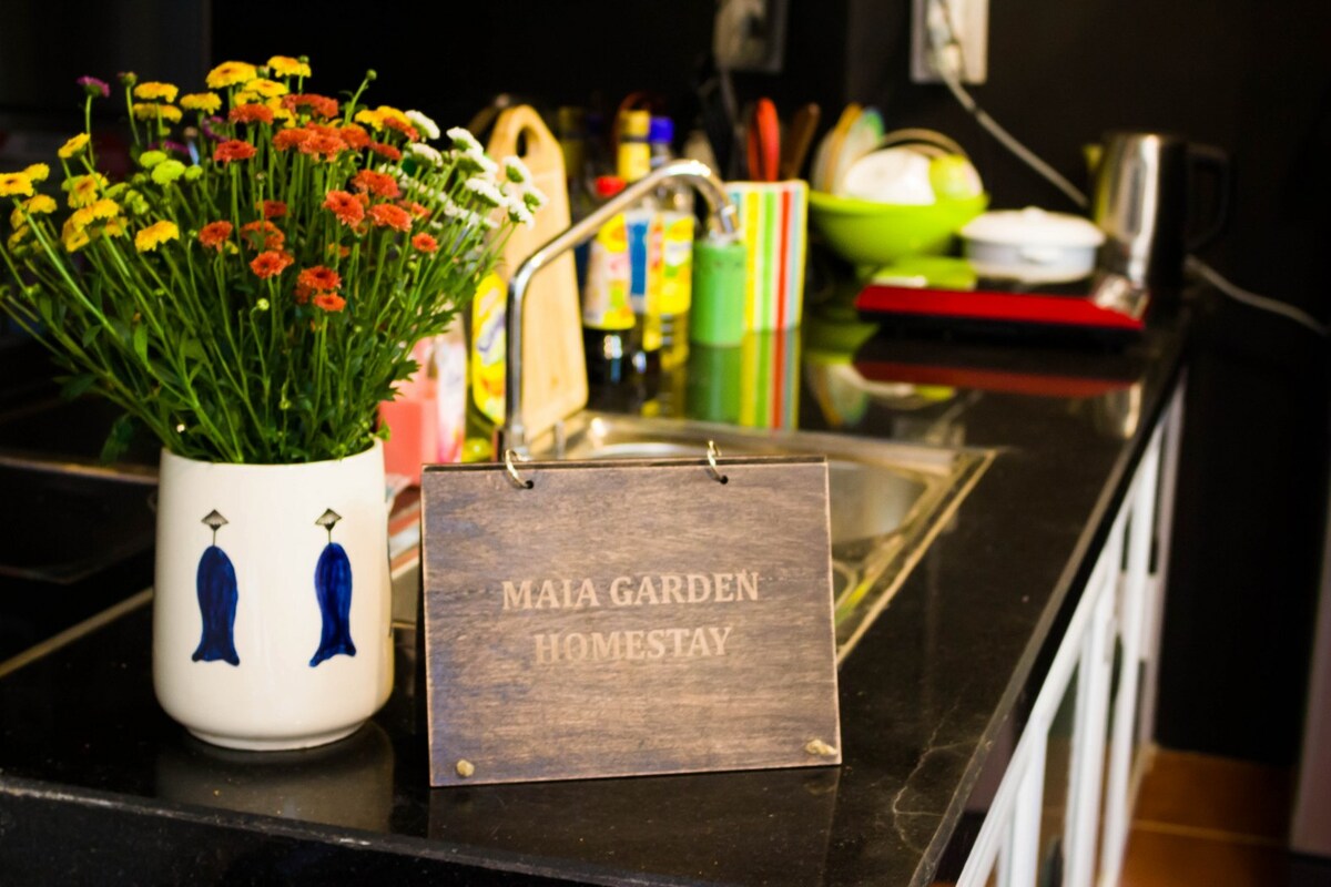 A kitchen space is depicted, featuring a dark countertop and a small sink area. A decorative sign reading 'MAIA GARDEN HOMESTAY' rests on the surface, accompanied by a bouquet of vibrant flowers in a white vase. Colorful kitchen items and utensils are visible in the background.